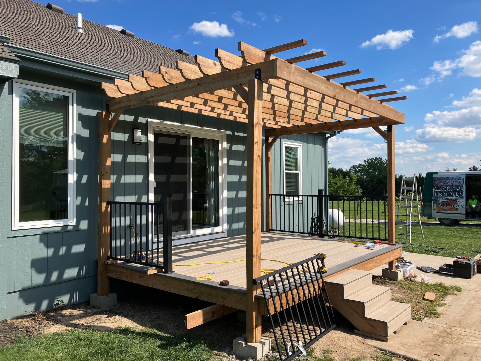 Wooden pergola over a deck attached to a blue house with black railings and stairs.