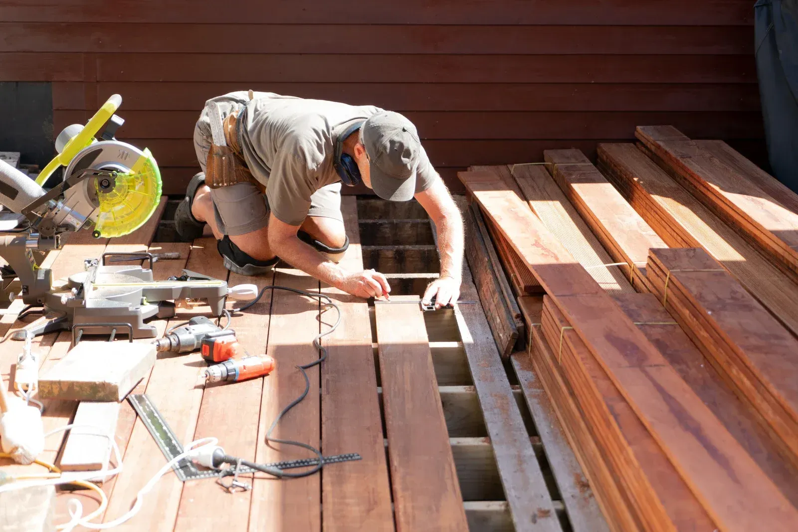 A man installing wooden deck boards, kneeling on the deck with tools and a saw in the outdoor sun.
