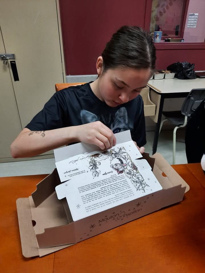 Une jeune fille est assise à une table et ouvre une boîte à pizza.