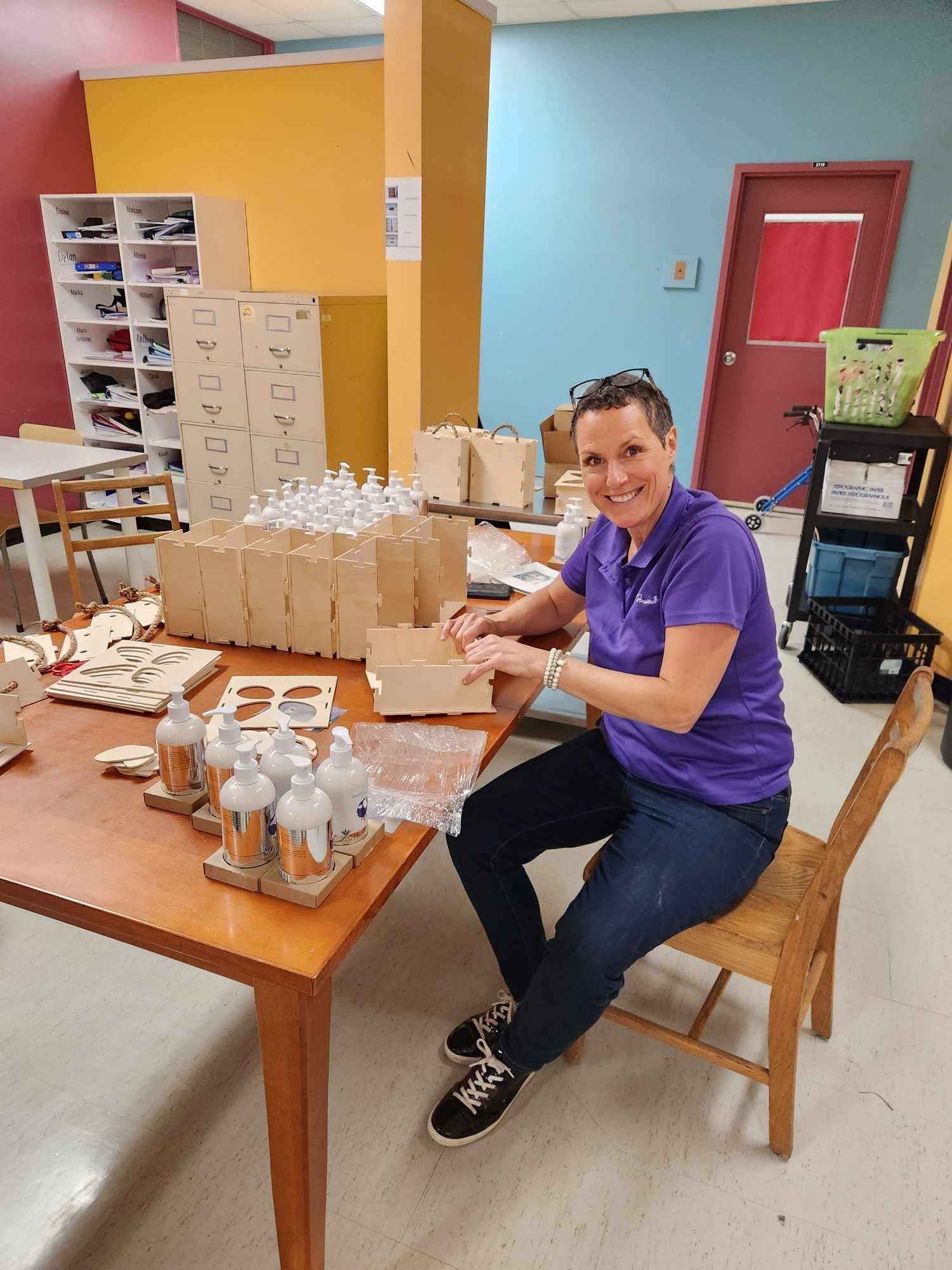 Une femme en chemise violette est assise à une table avec des bouteilles dessus.