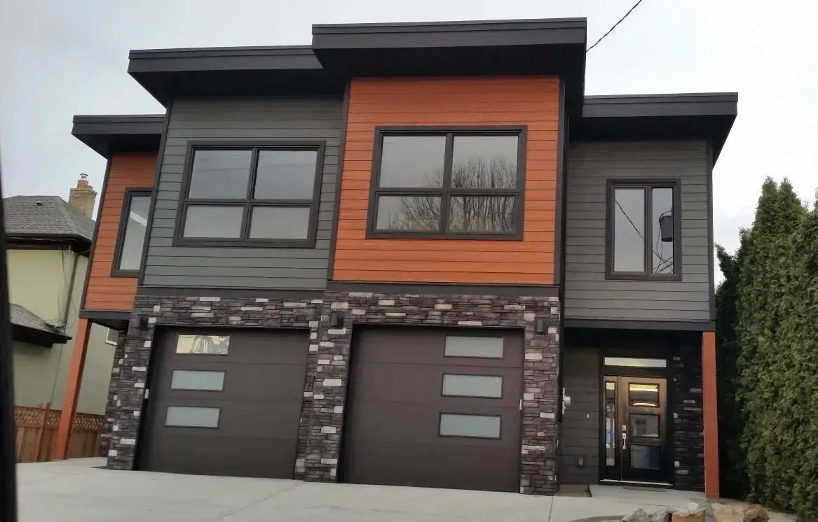 Modern two-story house with brown garage doors, gray and orange siding, stone accents, and large windows.