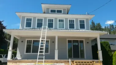 Two-story gray house with white trim, porch, and a ladder leaning against it on a sunny day.