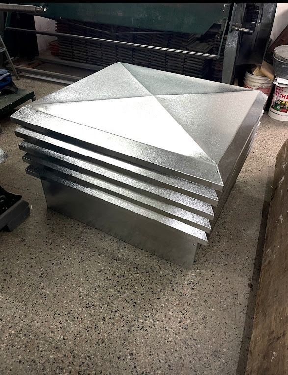 Rejillas de ventilación de techo de metal plateado apiladas con remates piramidales en el suelo de un taller.