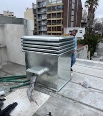 Caja de ventilación de climatización en la azotea con conductos metálicos en un tejado plano, con edificios de la ciudad al fondo.