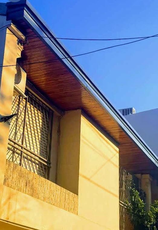Exterior soleado de una casa con paredes amarillas, un alero de madera y ventanas enrejadas contra un cielo azul.