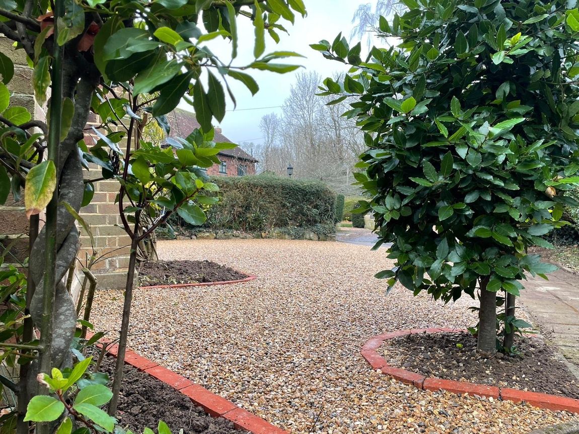A gravel driveway with trees in the foreground and a brick wall in the background.