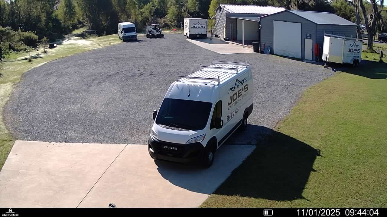 A white work van parked on a concrete driveway in front of a gravel lot with sheds and other vehicles in the background.