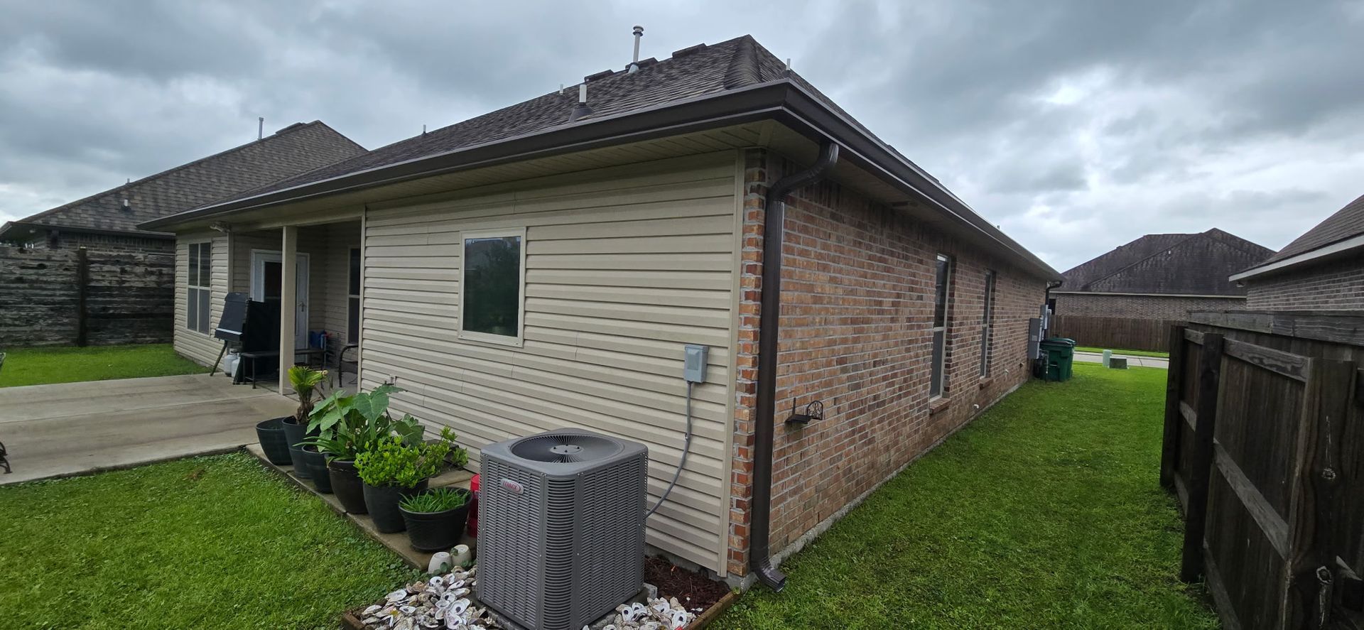 A side view of a house with beige vinyl siding and brick walls, featuring a backyard lawn and an AC unit.