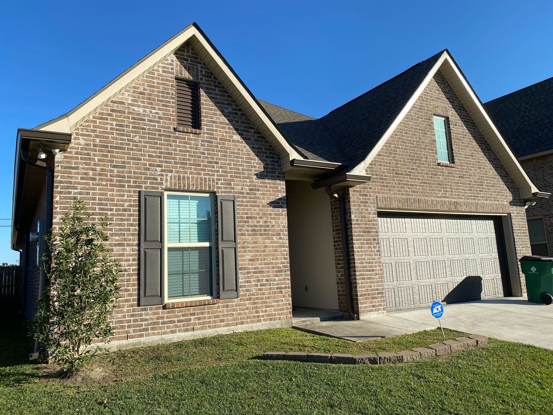 A brick house with a garage and a green trash can in front of it.