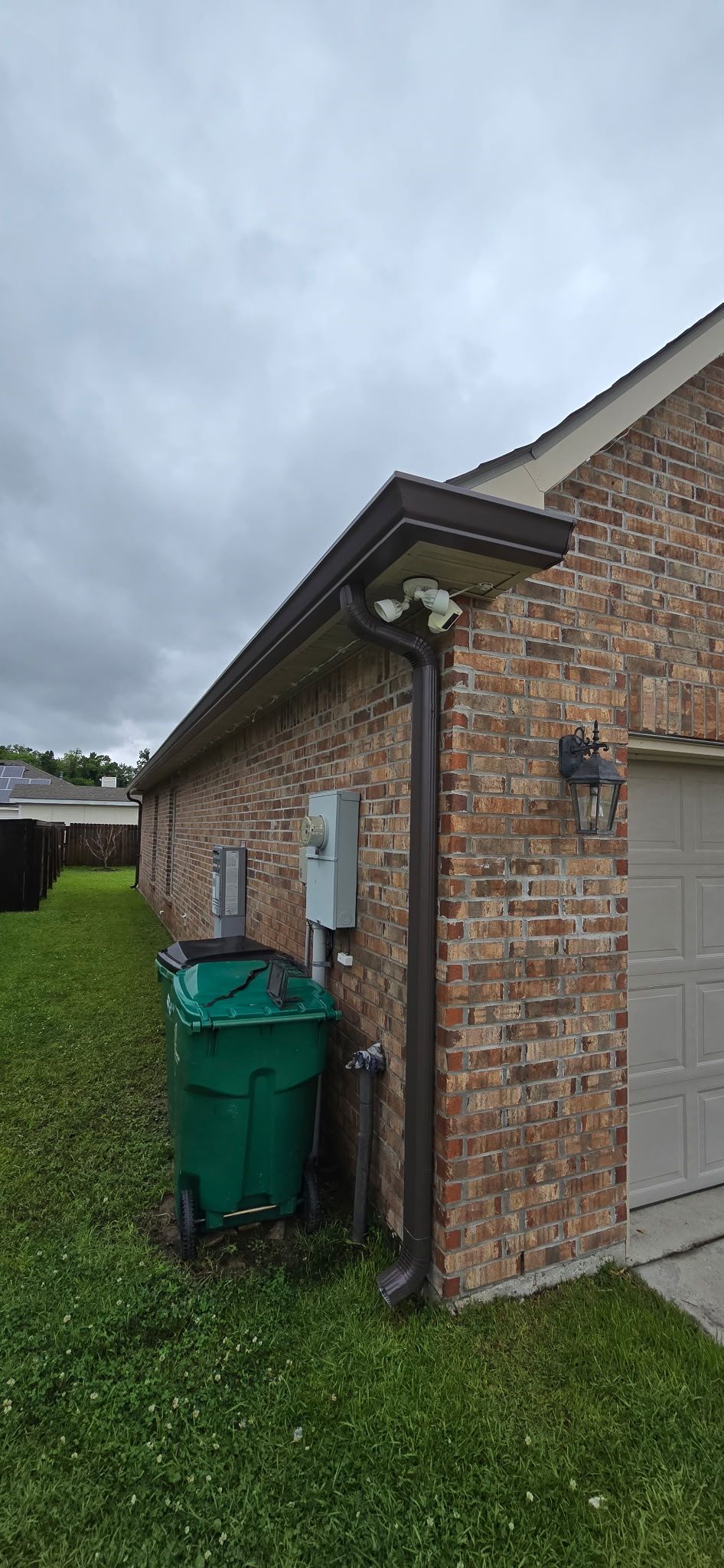 A green trash bin sits outside a brick house near a drainpipe and utility boxes under an overcast sky.