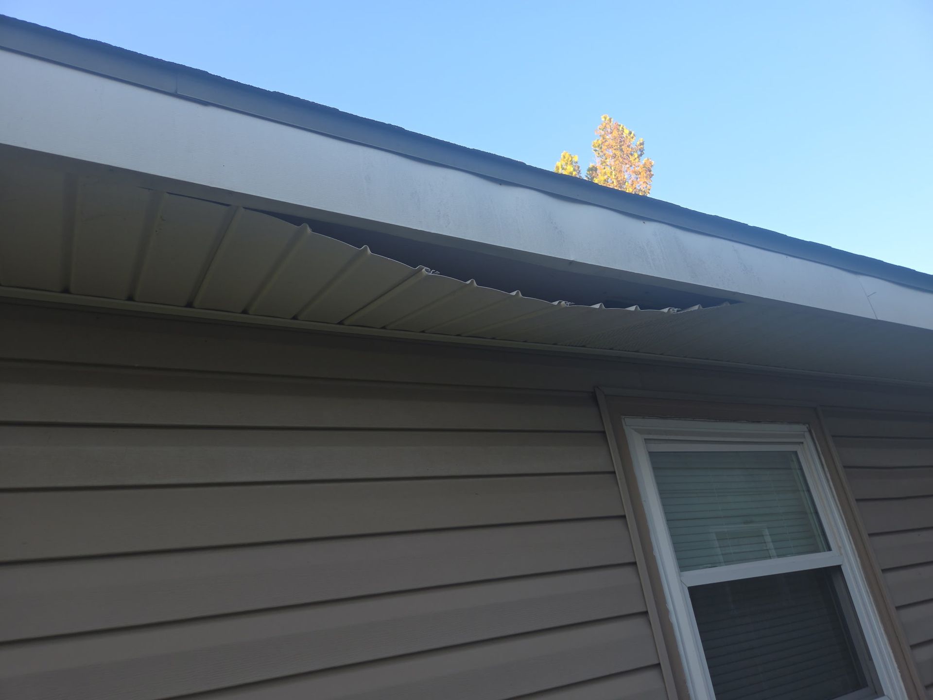 A close-up of a damaged, sagging soffit panel under the eaves of a house with light brown siding and a small window.
