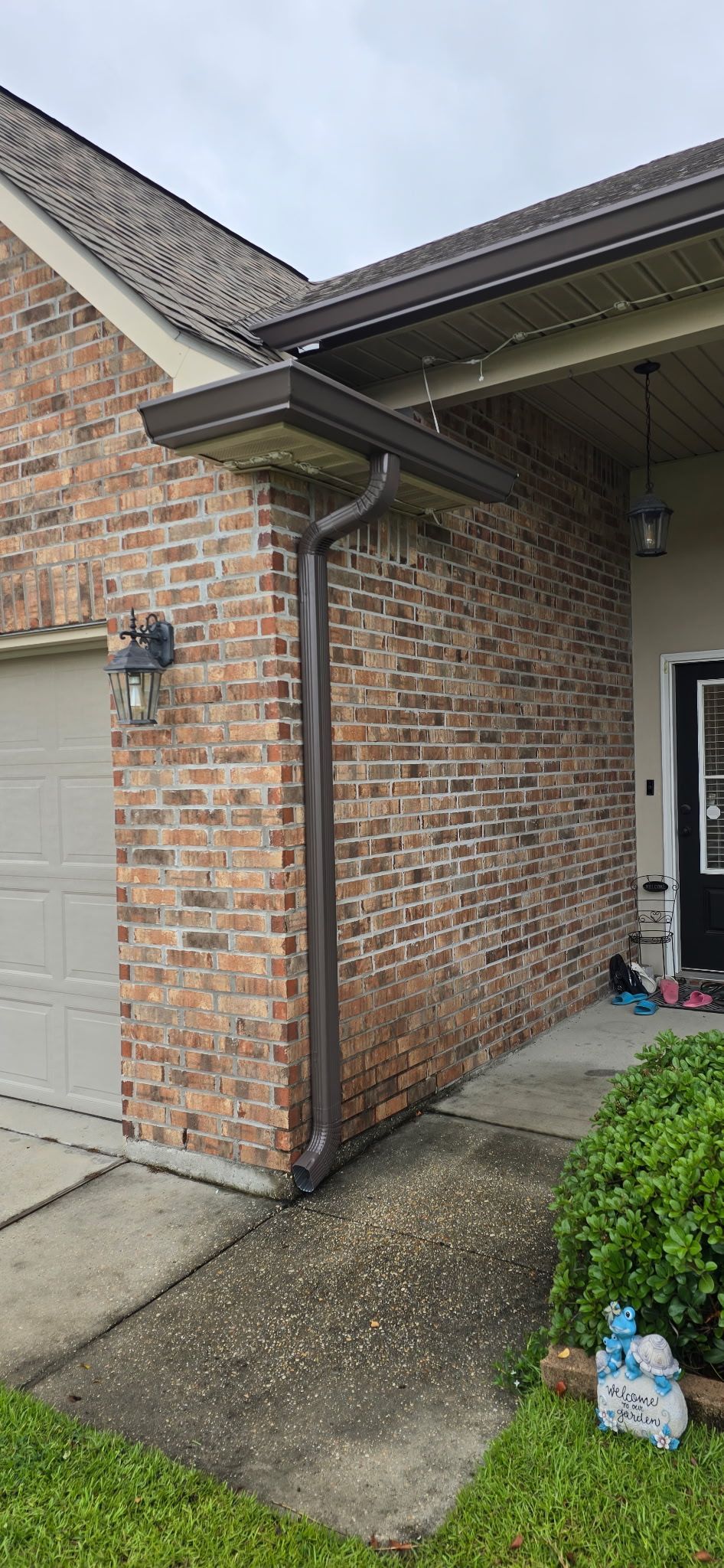 A dark brown gutter downspout mounted on the side of a brick house near a garage and concrete walkway.