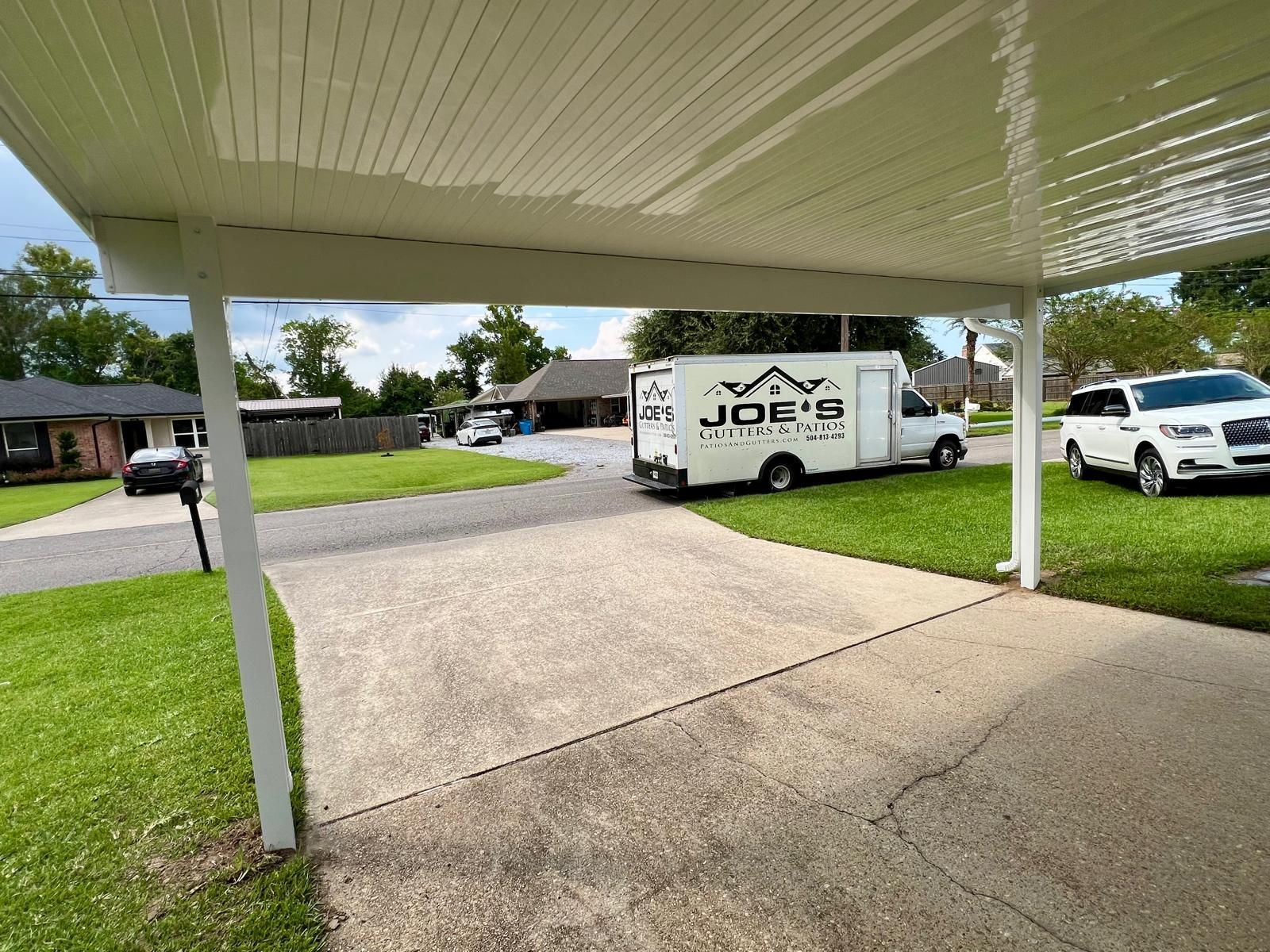 A driveway viewed from beneath a white carport, featuring a Joe's Moving truck and a white SUV parked on a sunny day.