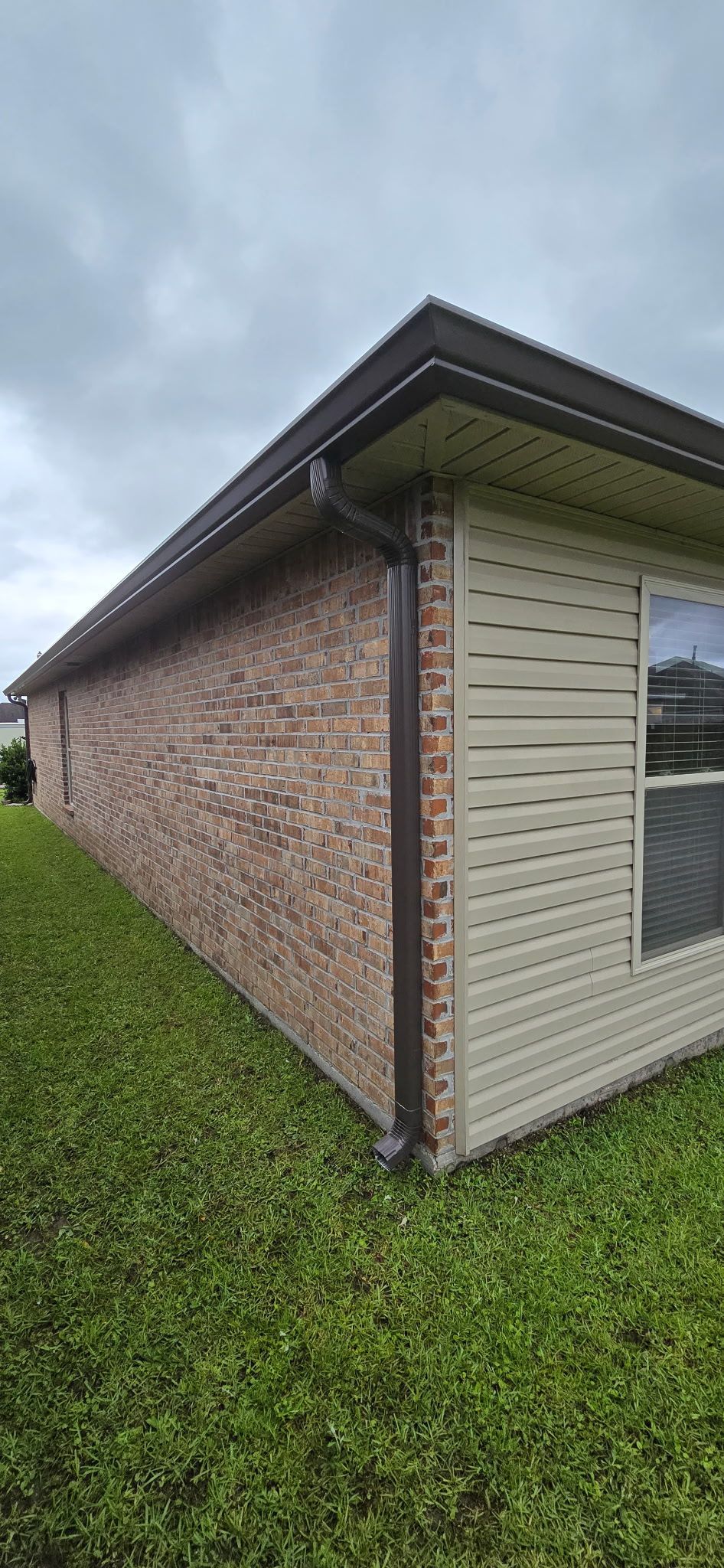 A side view of a house exterior featuring a brick wall meeting a section of tan horizontal siding under a roof overhang.