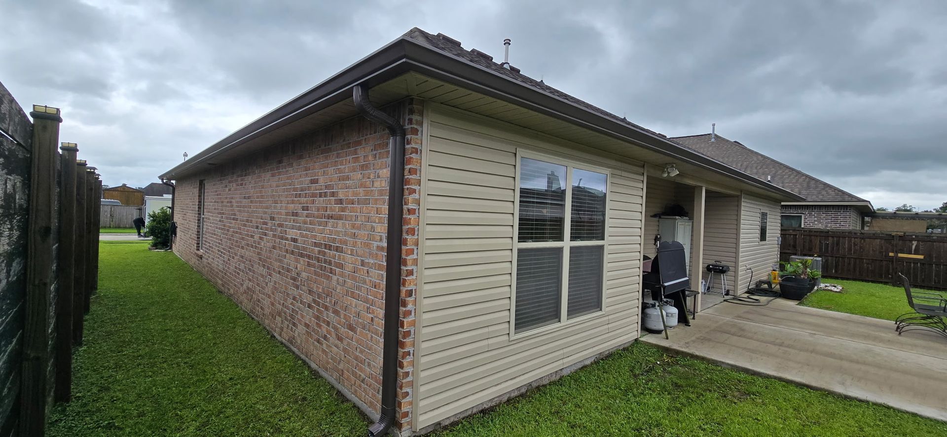 The side and corner of a brick house featuring beige horizontal siding, a square window, a driveway, and a grassy yard.