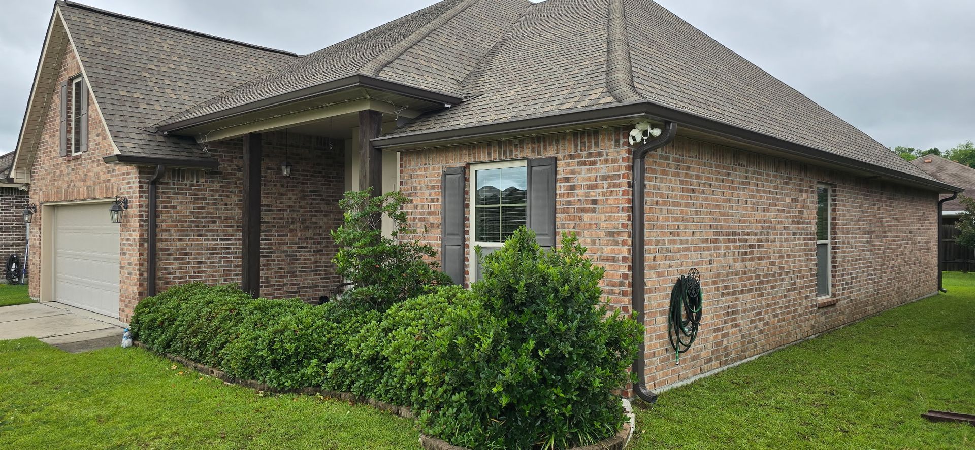 A single-story brick house with a front-facing garage, grey roof, and landscaped bushes in a residential yard.