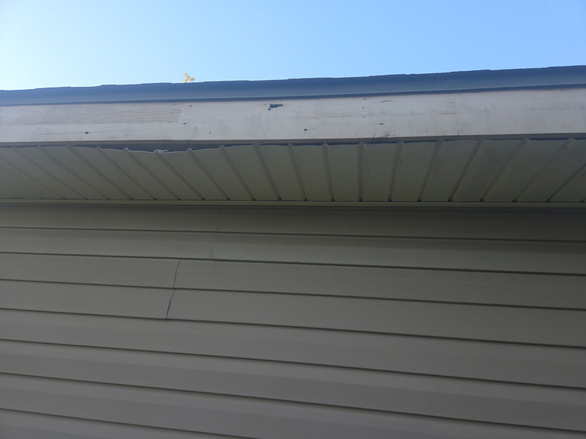 A low-angle view of a house exterior showing a detached and sagging soffit panel beneath the wooden roof fascia.