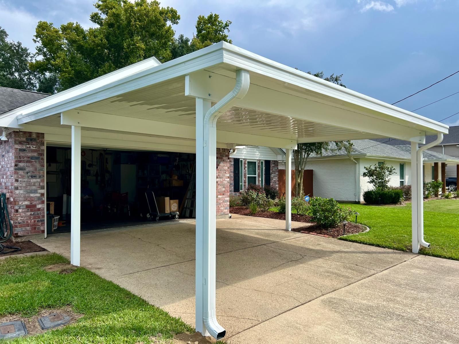 A white metal carport attached to a brick house next to a concrete driveway and green lawn under a partly cloudy sky.