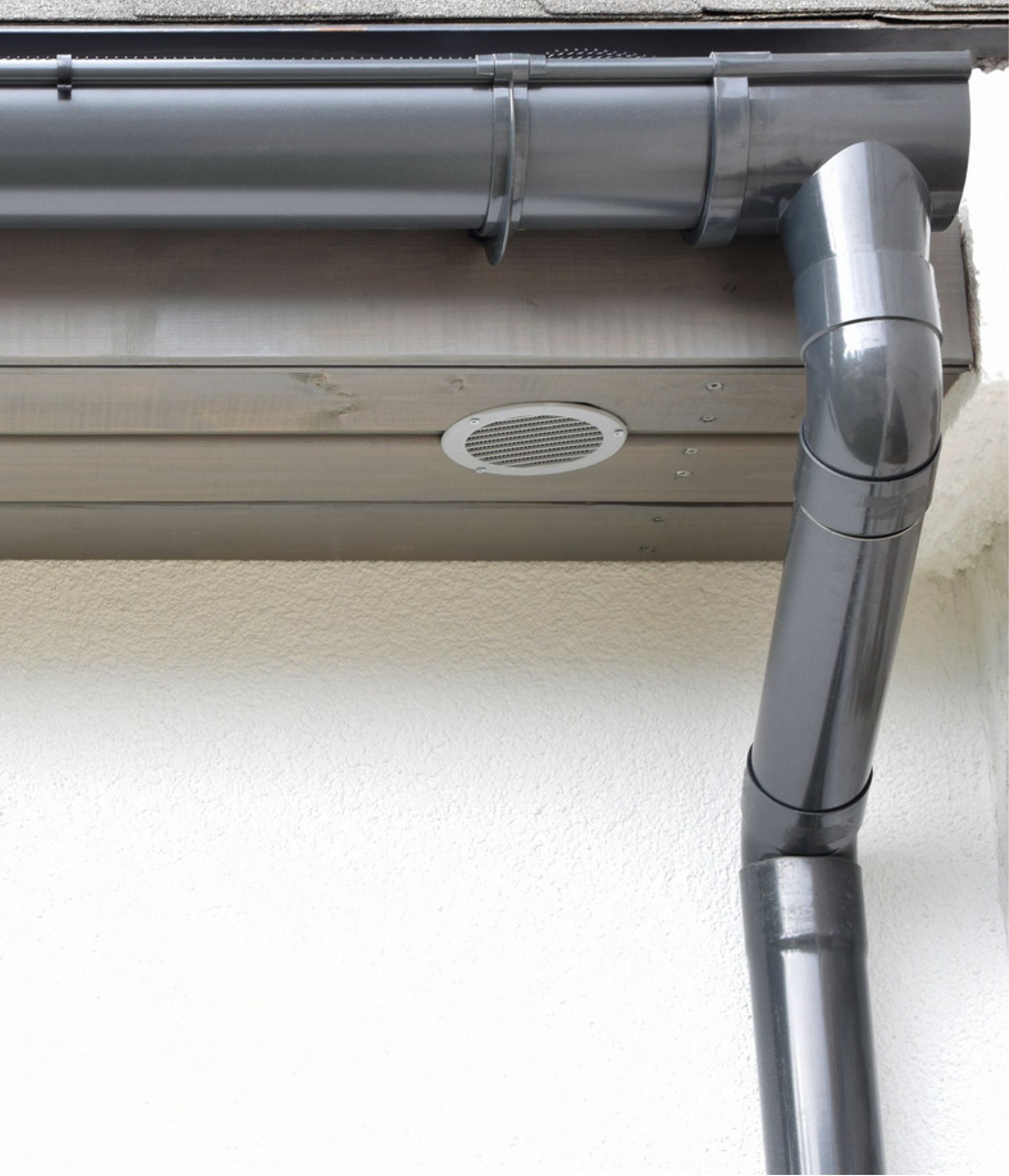 Gray rain gutter and downspout attached to a building's exterior, near a white wall and brown paneling.