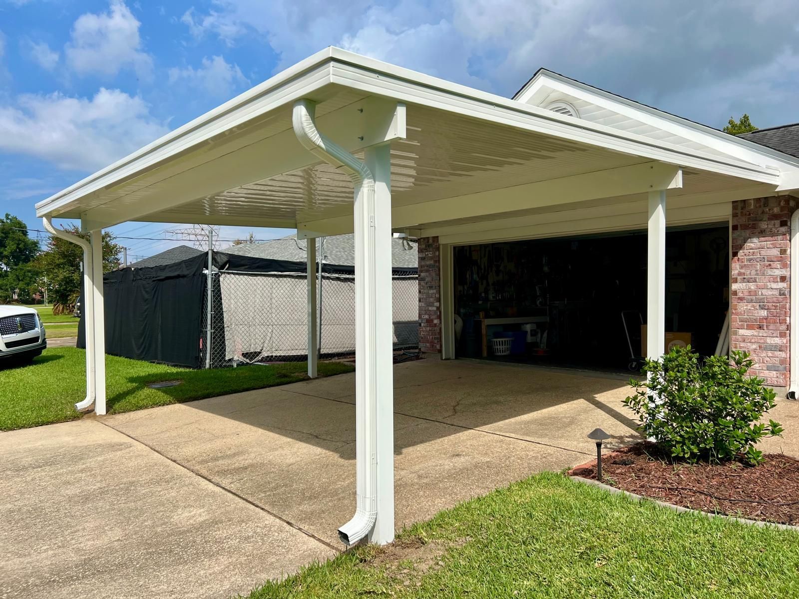 A white carport with a metal roof and downspouts attached to a brick house, overlooking a residential concrete driveway.