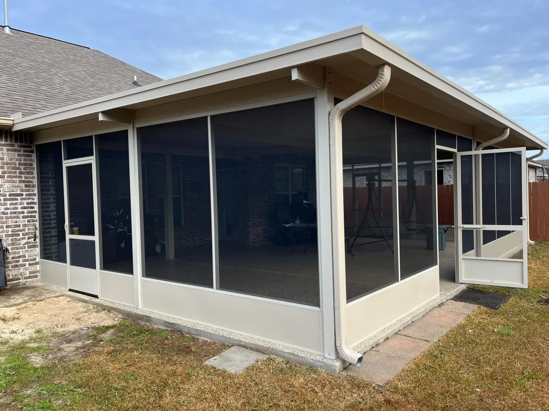 A beige, screened-in patio enclosure with two entry doors attached to the back of a brick house.