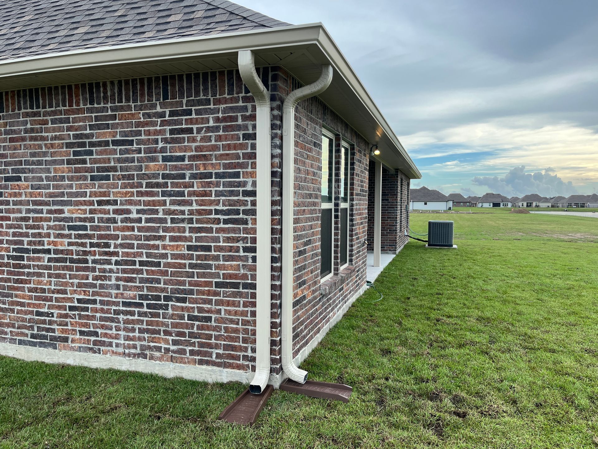 A corner view of a brick house with white downspouts extending onto brown metal splash blocks on a grassy lawn.