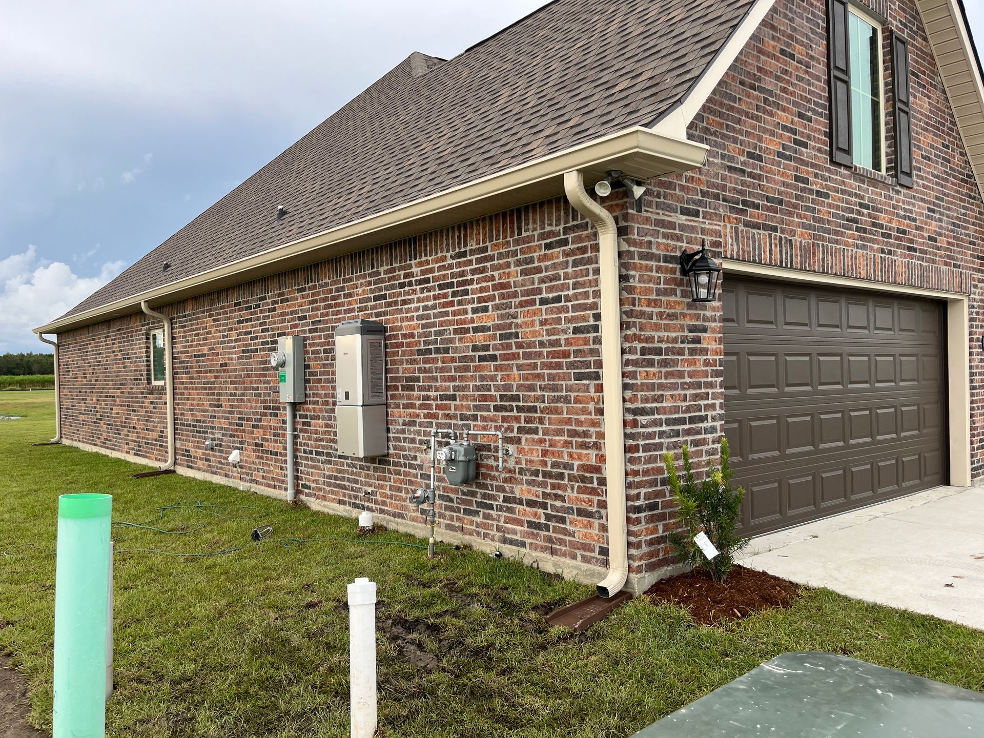 Side view of a brick house exterior with a garage, utility meters, and a green landscape under a cloudy sky.