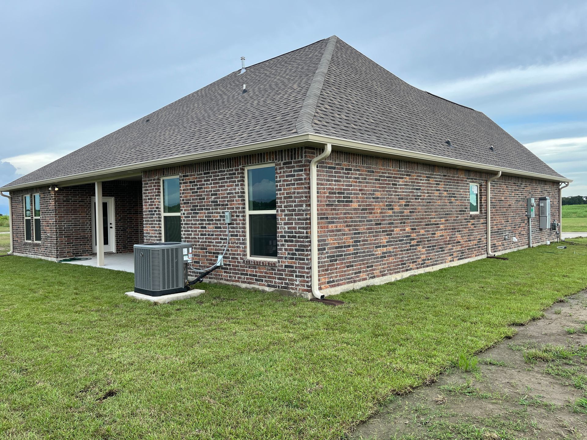 A side view of a modern, single-story brick house with a gray shingled roof, an air conditioning unit, and a grassy lawn.