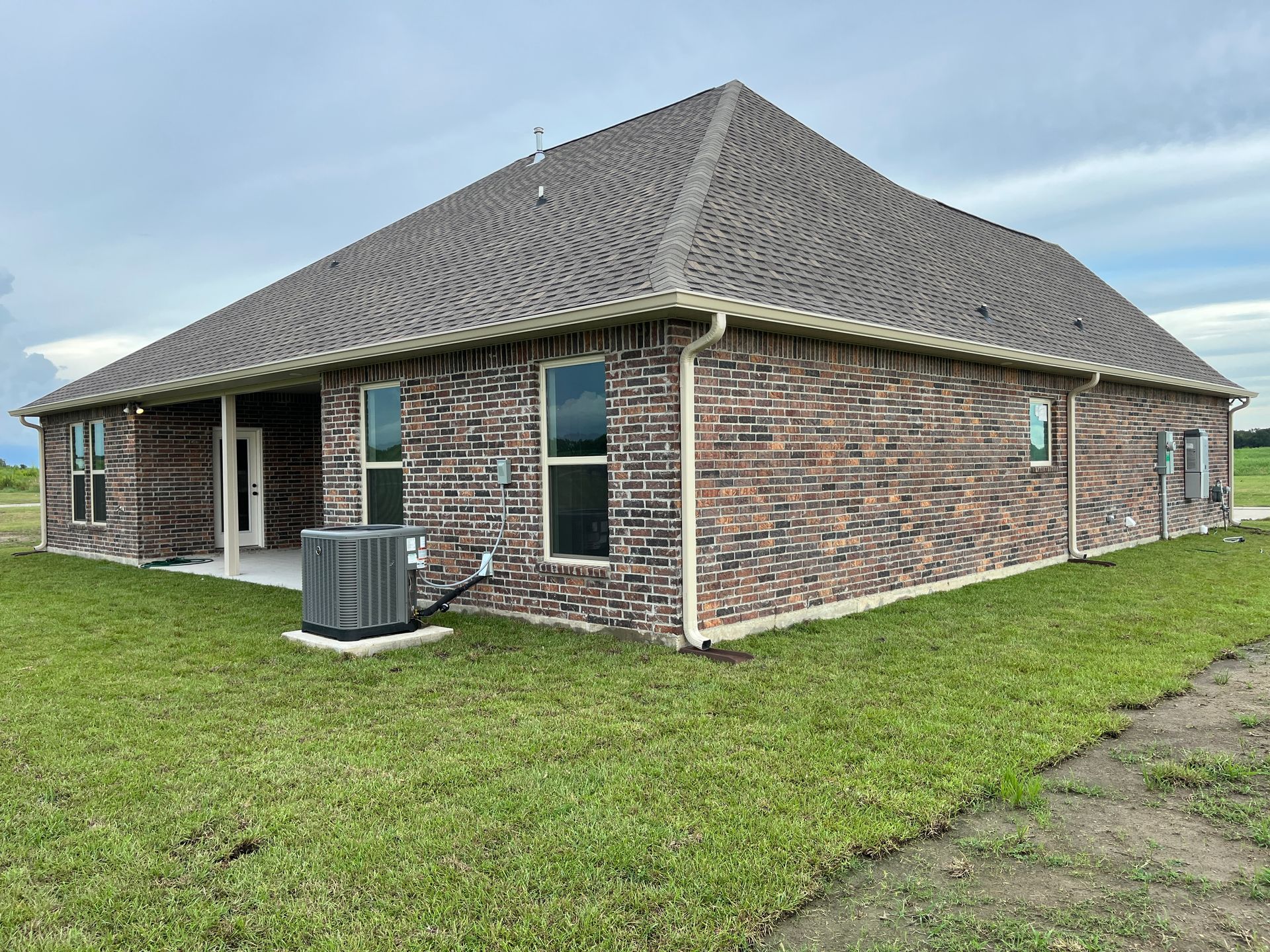 A rear view of a single-story brick house with a dark shingled hip roof and an outdoor HVAC unit on a grassy lawn.