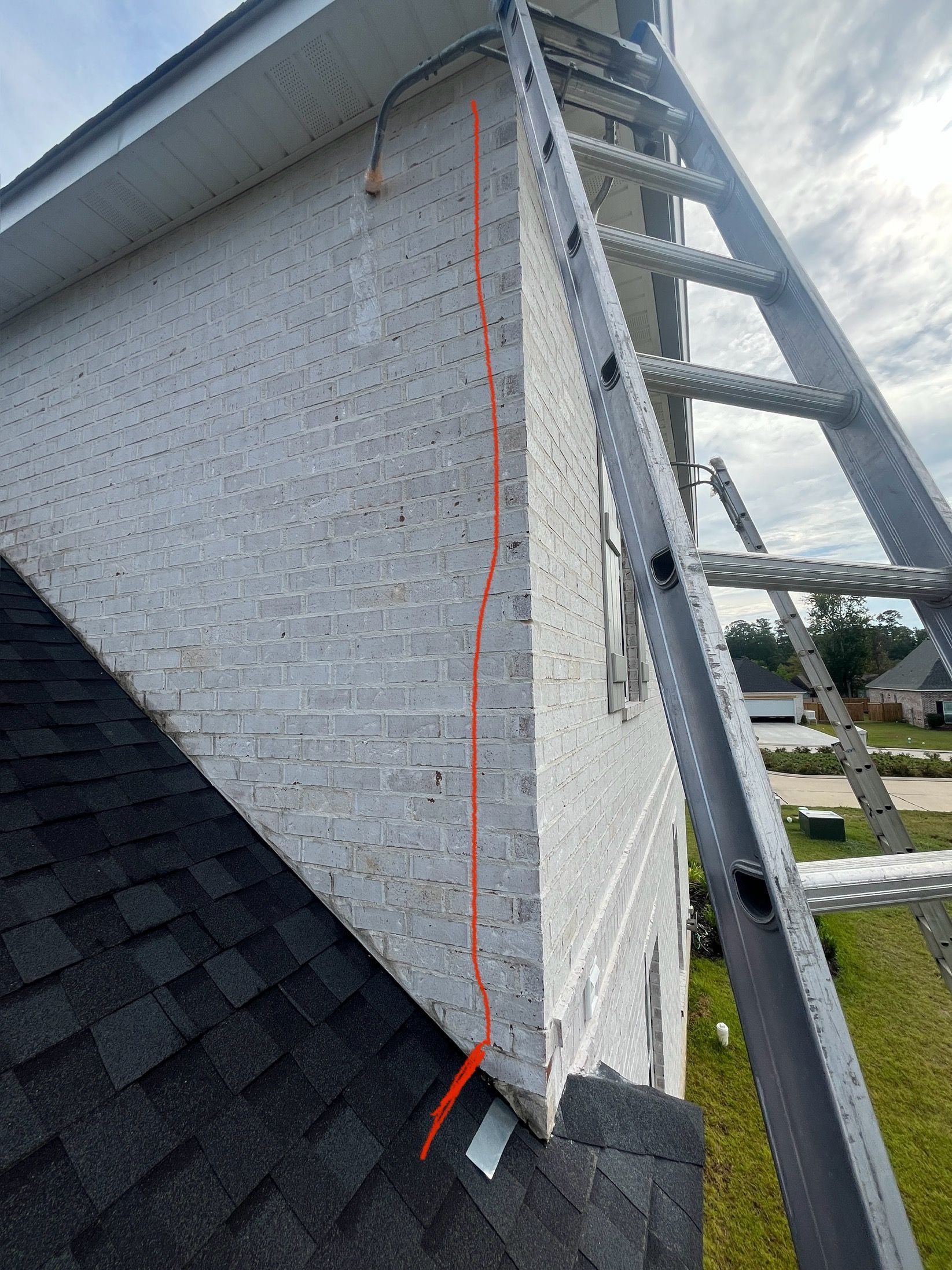 A view from a ladder looking at a white brick wall with a vertical red line marking a crack above a roofline.