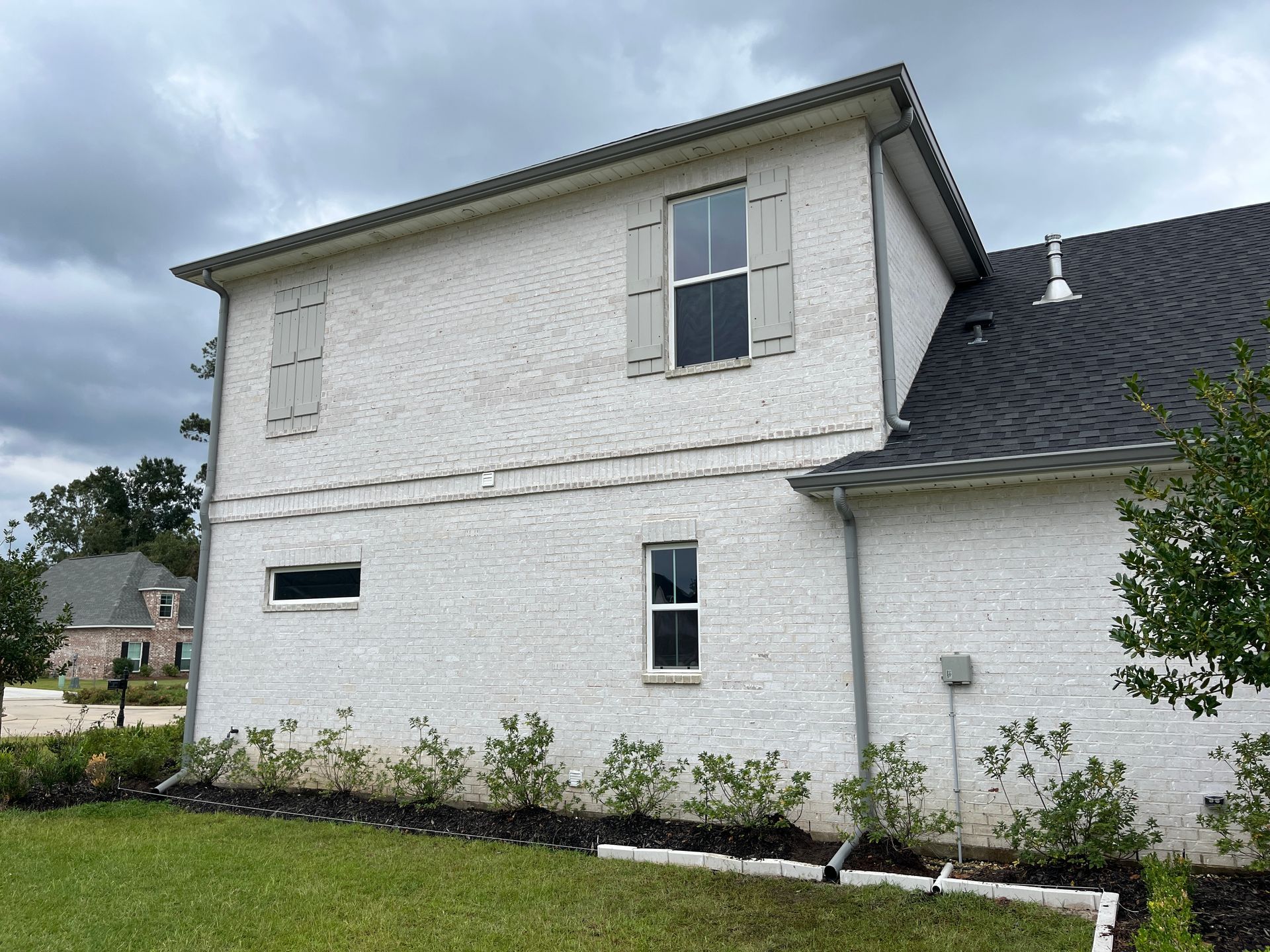 A side view of a white brick, two-story house with gray shutters and a dark shingled roof under a cloudy sky.