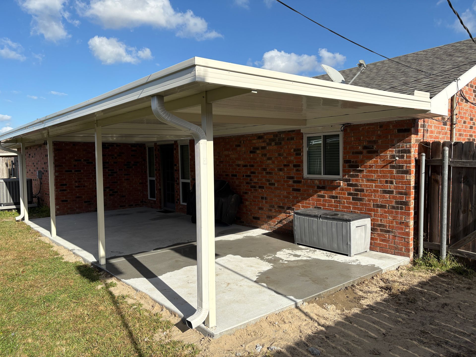 A brick house exterior with a large covered concrete patio, white support posts, and a plastic storage bin.