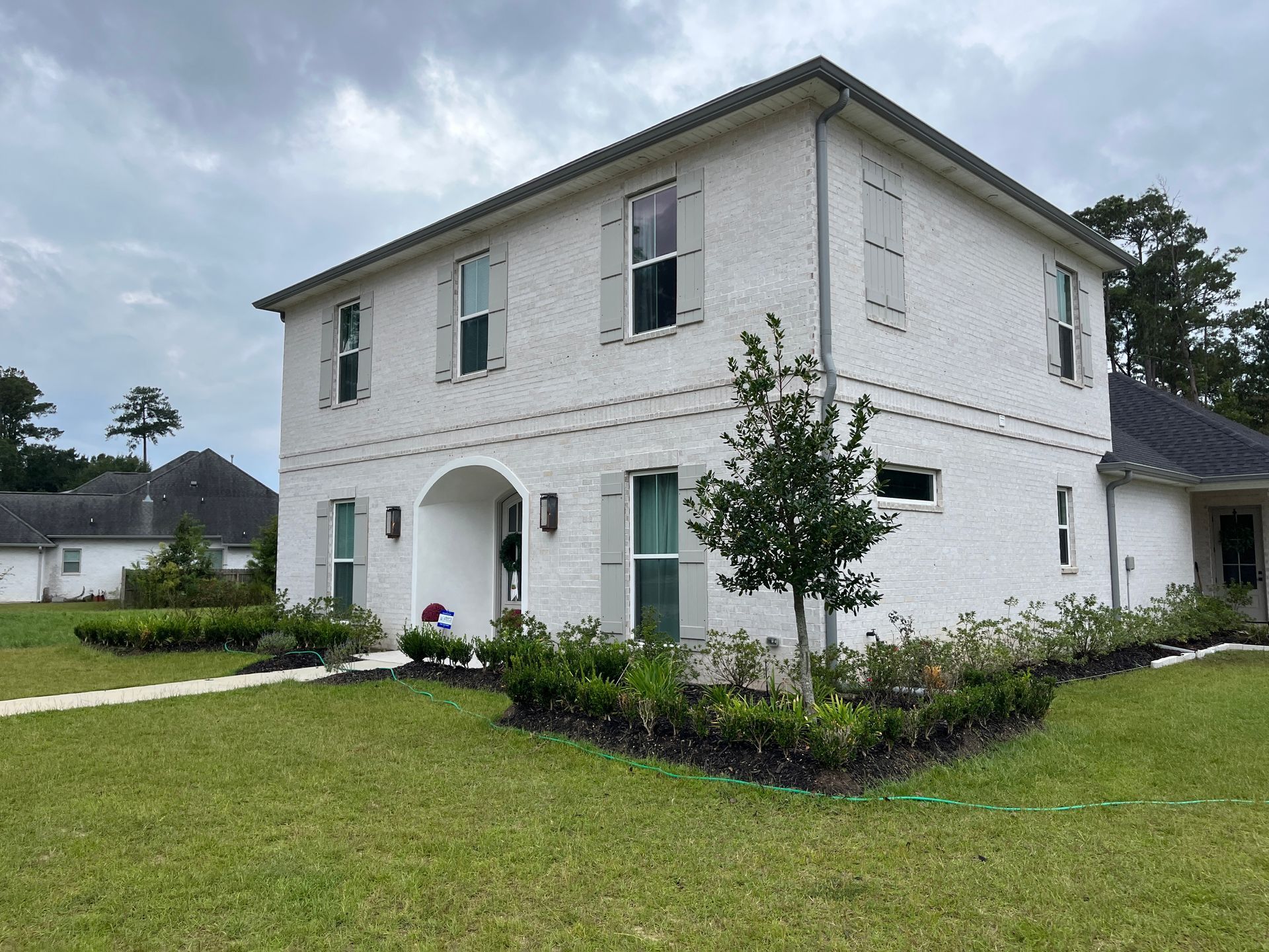 A two-story white brick house with gray shutters, a small front yard, and landscaping on a cloudy day.