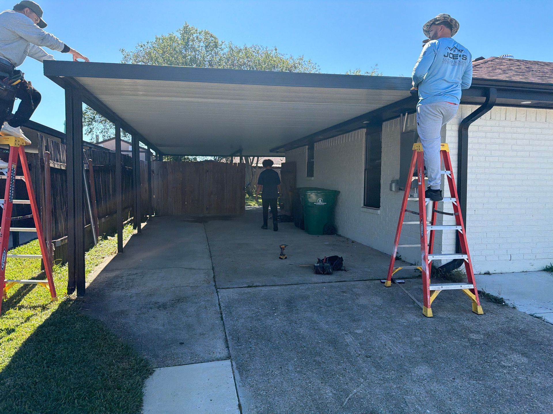 Three workers on ladders install a metal patio cover attached to the side of a brick house.