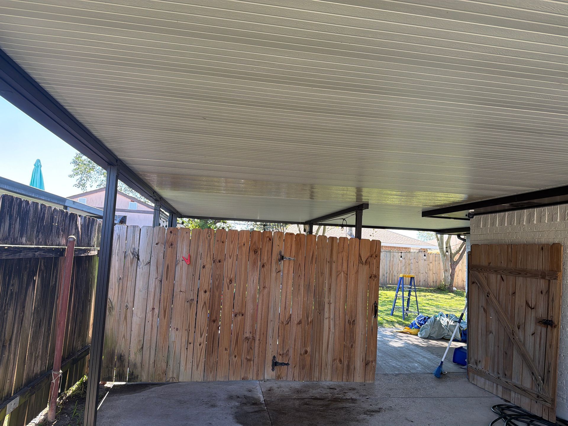 A wooden gate hangs mid-air under a covered carport, with a backyard visible in the background.