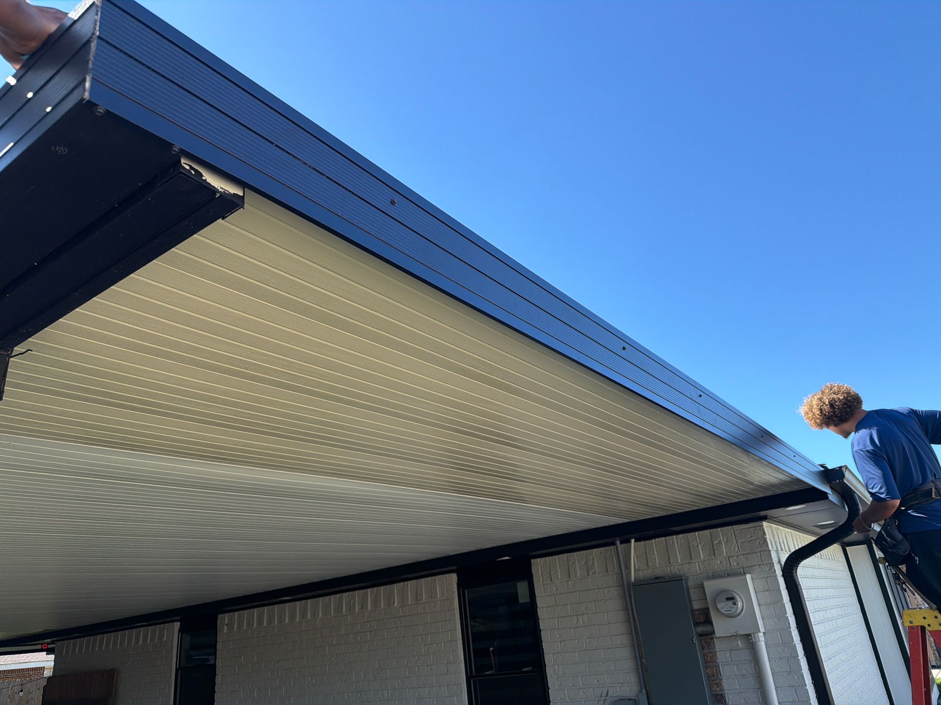A person on a ladder installs black trim along the edge of a tan slatted patio roof attached to a brick house.
