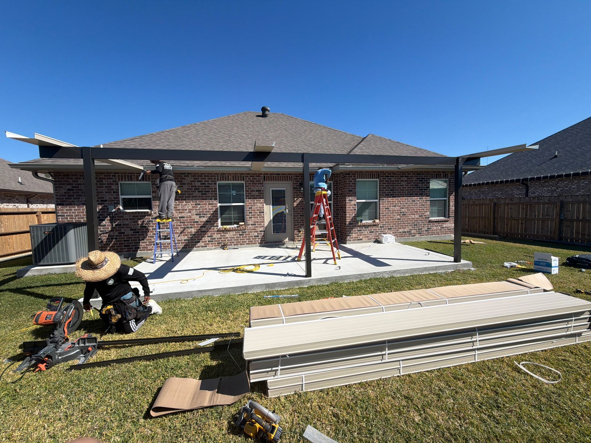 Workers assemble a black metal pergola frame on a concrete patio in a residential backyard on a sunny day.