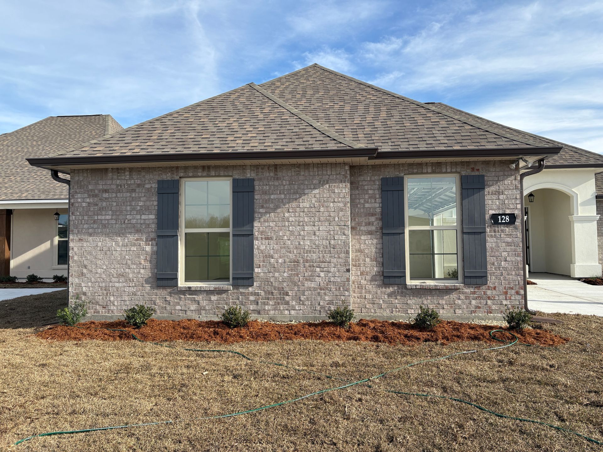 Single-story house with tan brick, a dark shingled roof, dark shutters flanking two windows, and fresh mulch in front.