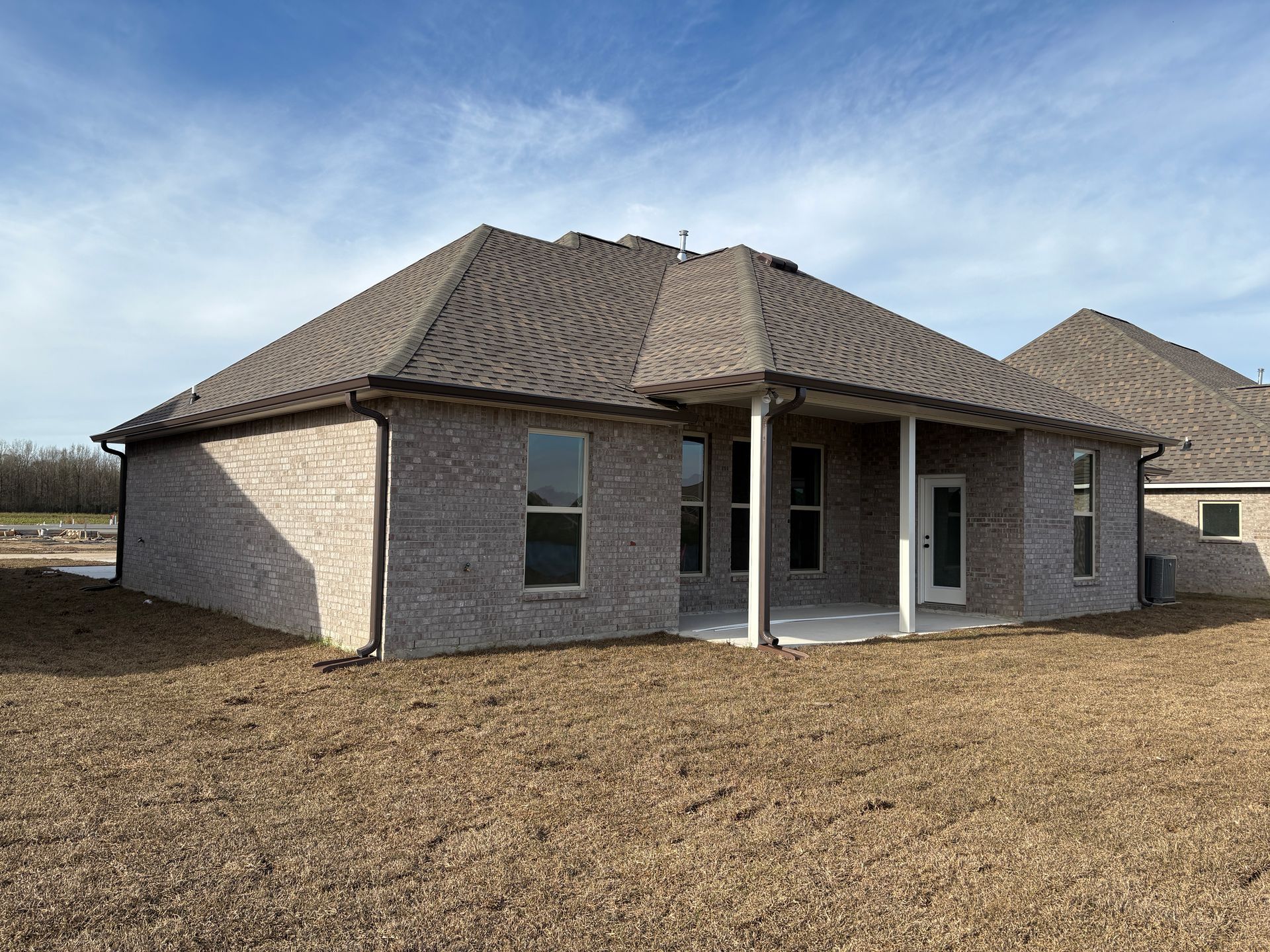 The back exterior of a new beige brick house with a covered patio and a large, dirt-covered backyard under a blue sky.