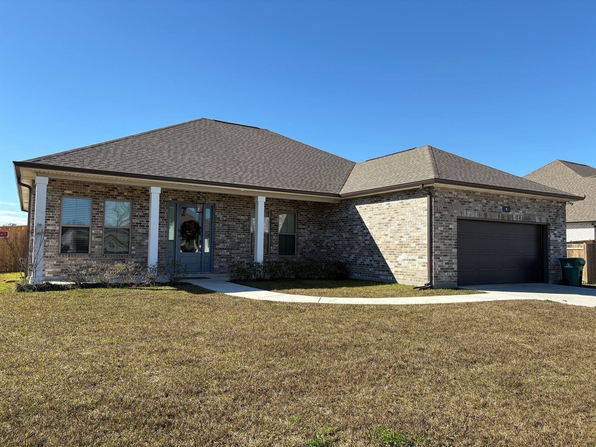 A single-story, tan brick house with a dark gray shingled roof, a covered front porch, and an attached two-car garage.