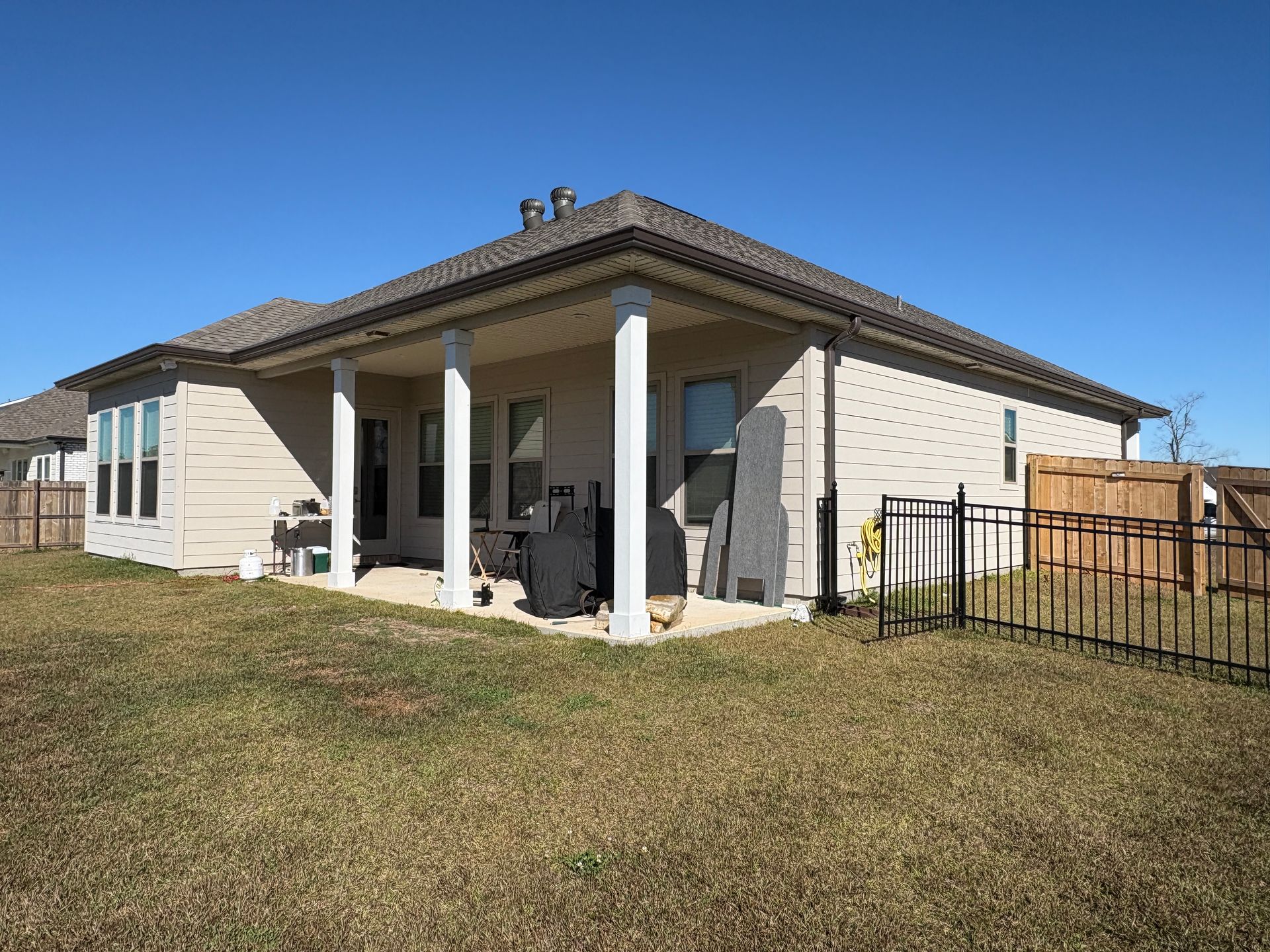 A tan-colored suburban home with a covered back patio, white pillars, and a metal fenced yard under a clear blue sky.