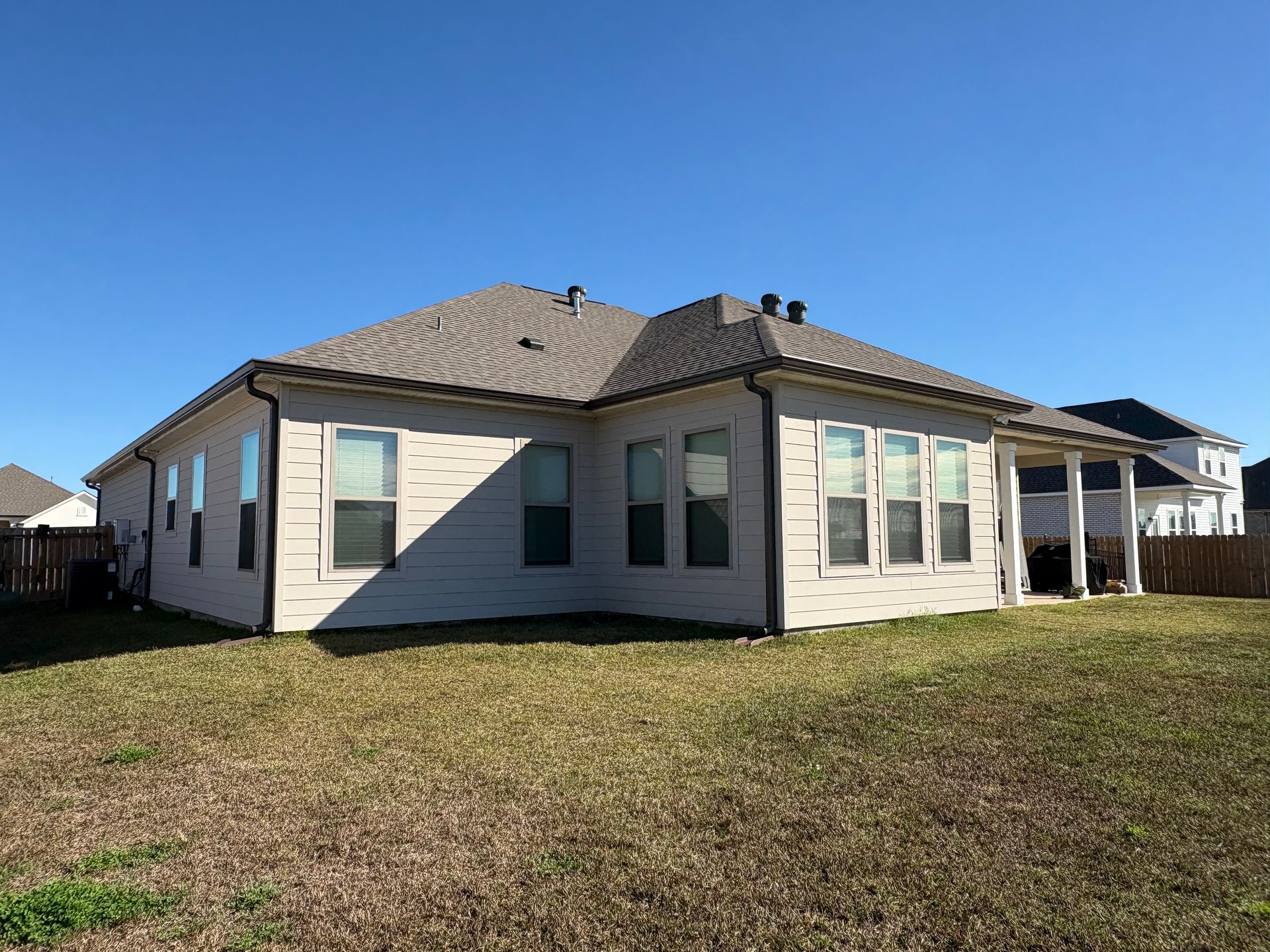 Backyard view of a beige single-story house with a tan shingled roof, large windows, and a covered patio on a sunny day.