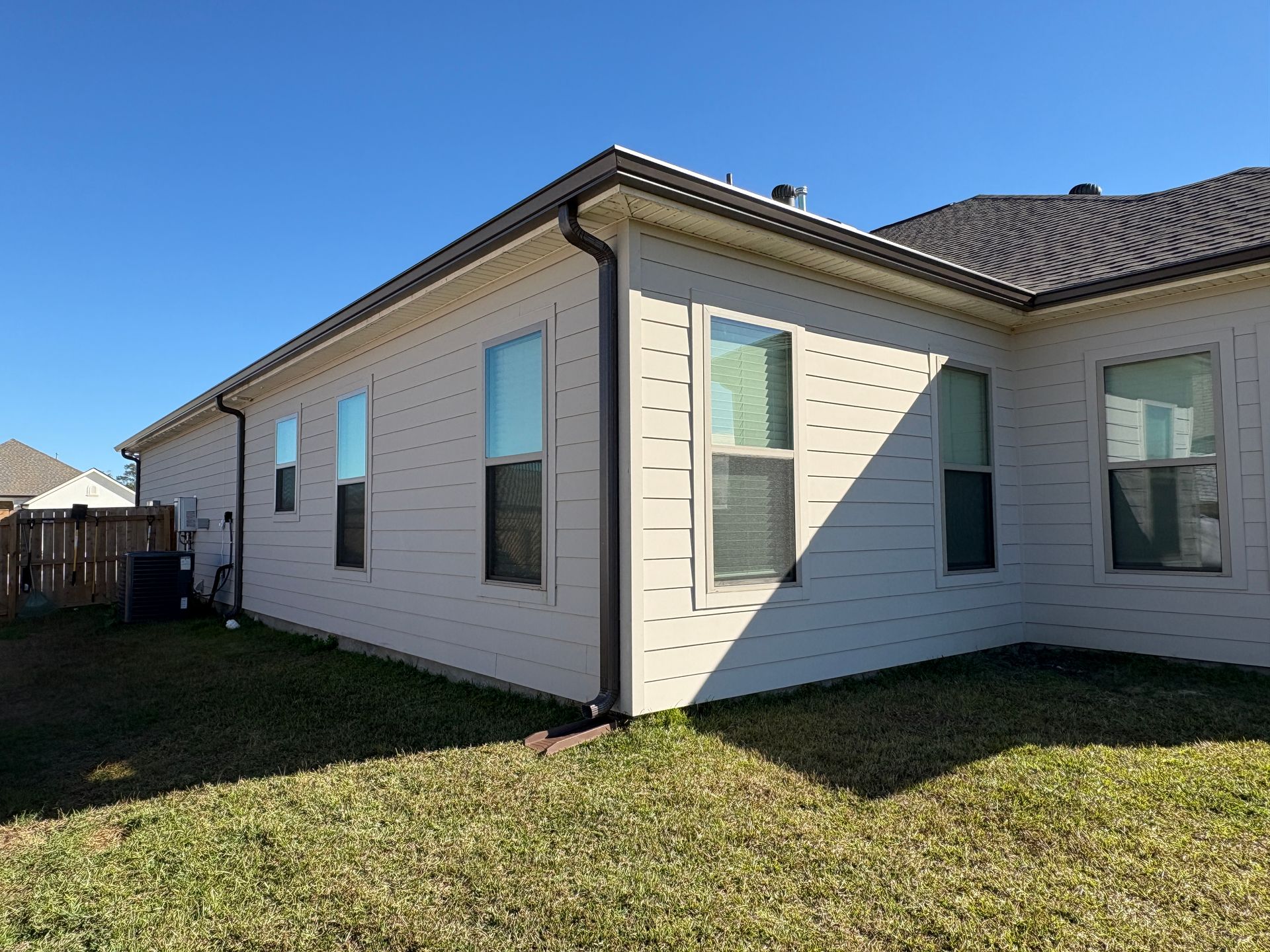 The side and back corner of a light-colored, single-story house with multiple windows and a brown gutter on a sunny day.