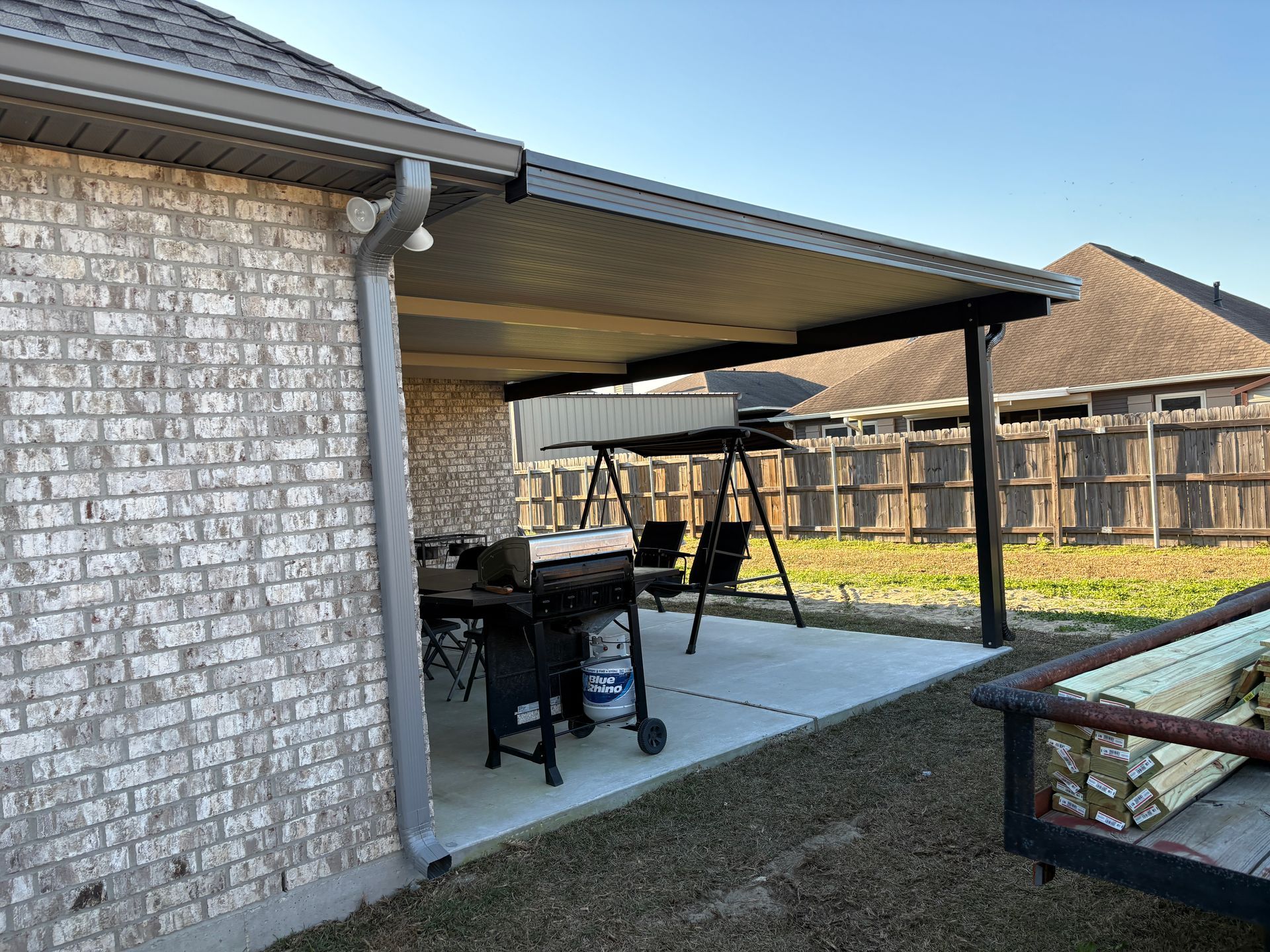 A covered patio with a charcoal grill and a swing set on a concrete slab against a brick house wall.
