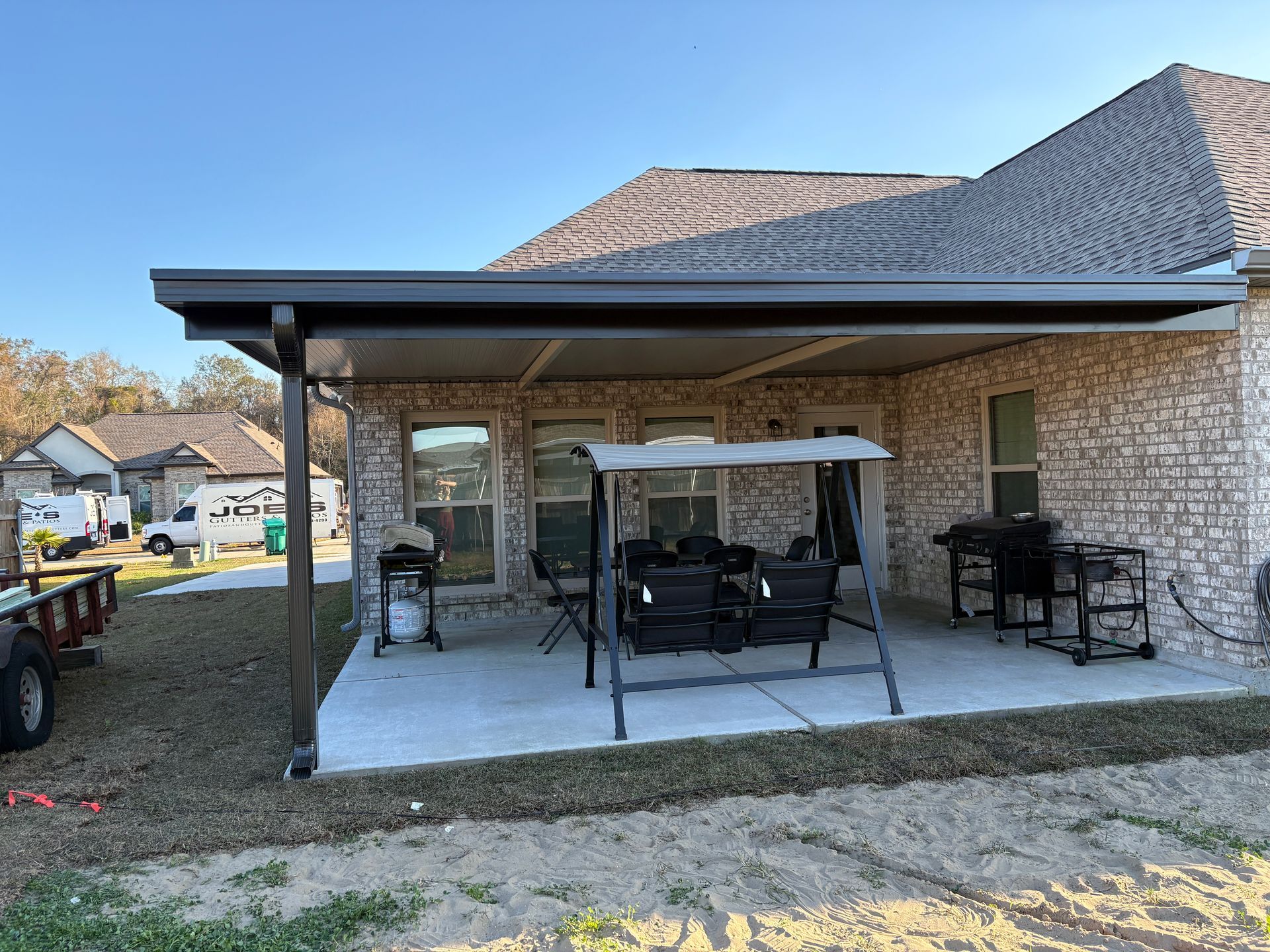 A concrete patio with a dark metal awning, outdoor seating, a grill, and a smoker attached to a brick house.