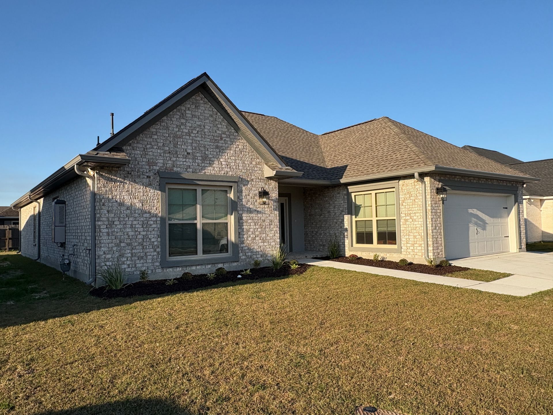 A single-story, light-colored brick house with a dark shingled roof, a front porch, and a two-car garage at sunset.