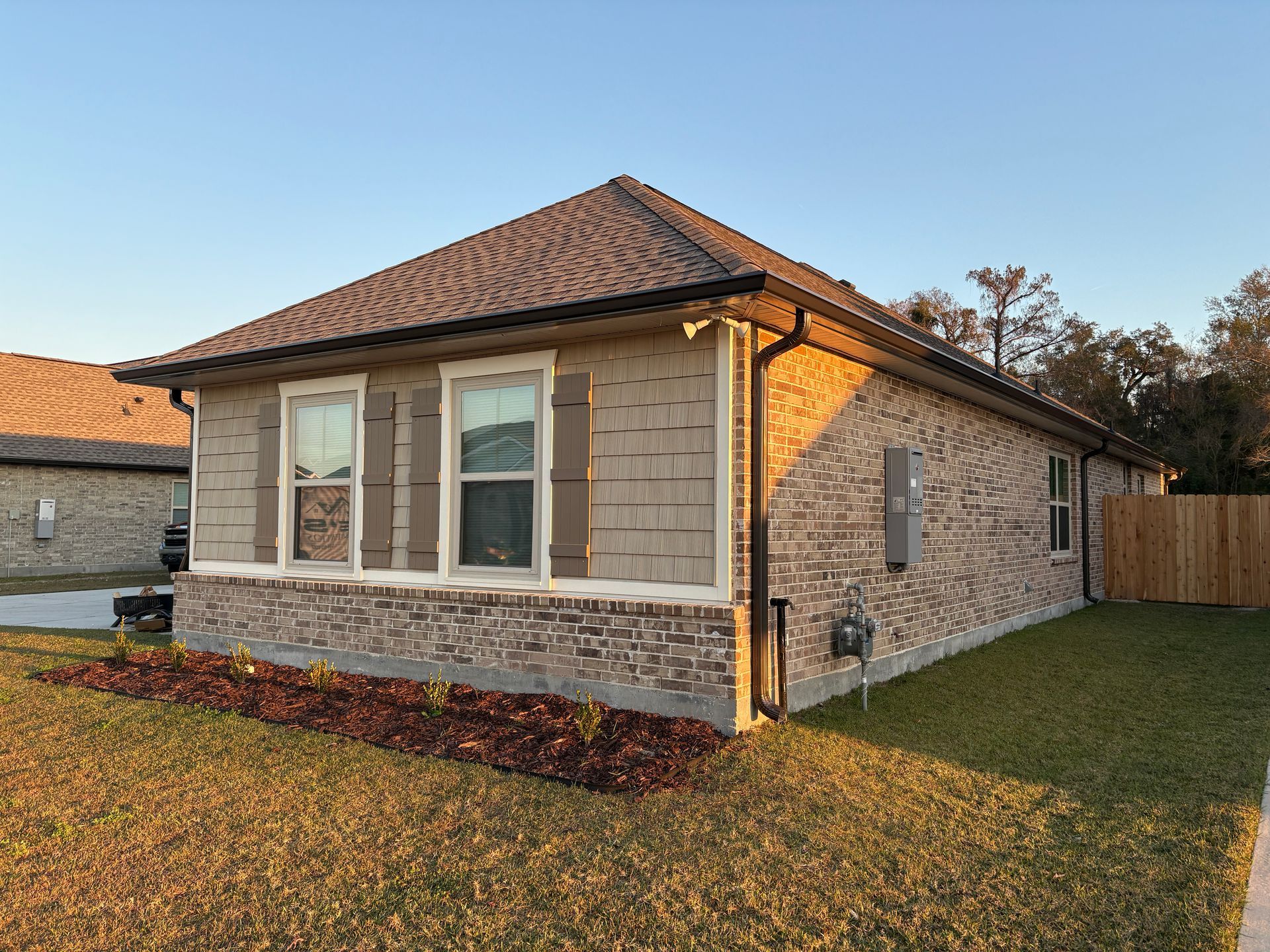 A corner view of a beige, single-story house with a stone base and dark shingles during the golden hour.