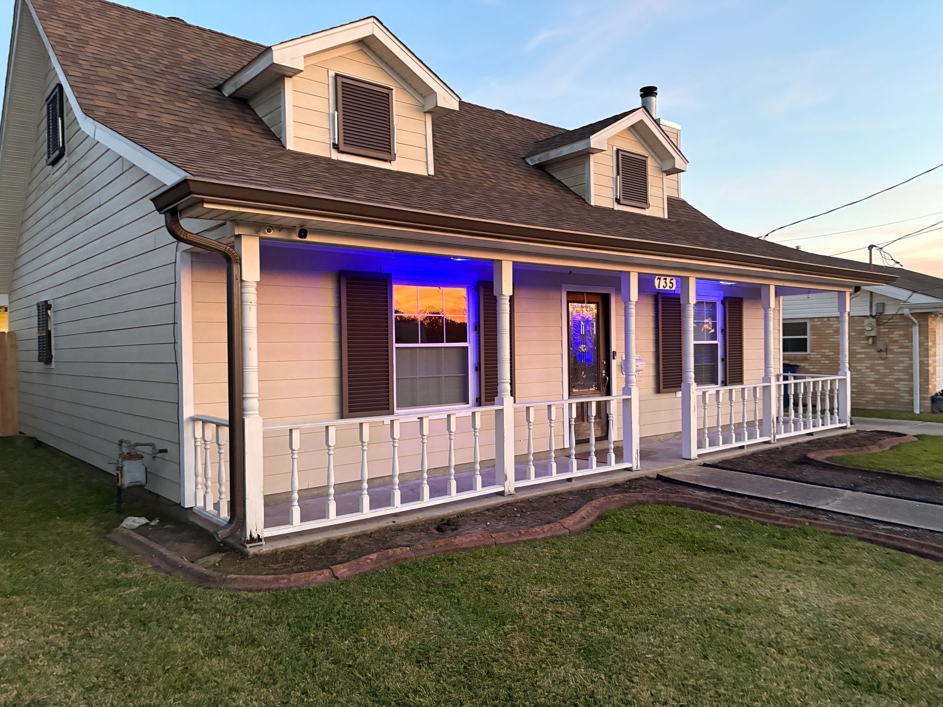 A tan, two-story house with a white front porch, brown shutters, and glowing blue lights reflecting in the front windows.