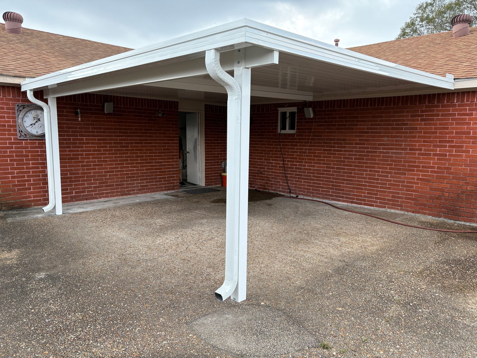 A white metal carport attached to a red brick house with an exposed aggregate concrete driveway.