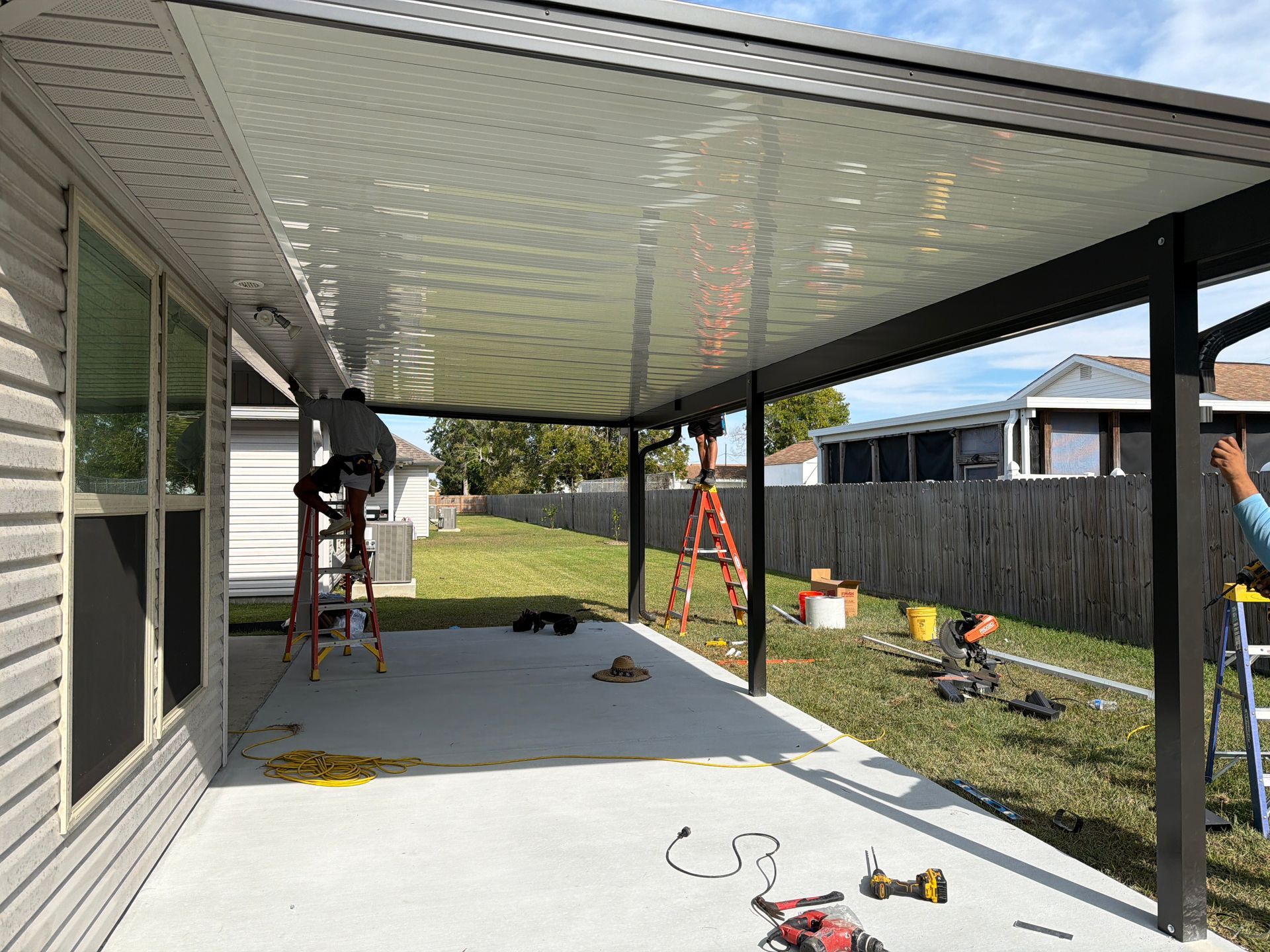 Workers install a white patio cover over a concrete slab on a sunny day.